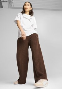 Maglietta bianca con logo Puma grigio, abbinata a pantaloni sportivi larghi marroni con cuciture laterali e un piccolo logo Puma bianco, indossati con sneakers.