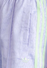 Lila träningsbyxor med ett subtilt blommönster, som har ljusgröna sidsömmar och en broderad logotyp på låret.