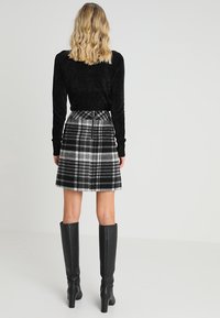 Schwarzes Langarmoberteil, karierten grau-schwarzen Schlagrock, knielange schwarze Lederstiefel, alles vor einem neutralen Hintergrund.
