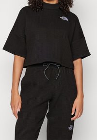 Svart topp med våffelstruktur, kortärmad och med hög halsringning, med en kontrasterande vit logotyp. Matchas med svarta byxor.