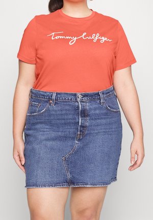 Kobieta w pomarańczowej koszulce Tommy Hilfiger i niebieskiej jeansowej spódnicy z przednim szwem i kieszeniami, stojąca na jednolitym tle.