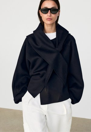 Kvinde iført en oversized sort slå-om-frakke, hvide bukser og sorte cat-eye solbriller står foran en ensfarvet baggrund.