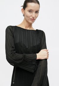 Vrouw draagt een doorschijnende zwarte jurk met lange mouwen en verticale strepen, raakt haar linkerarm aan, tegen een effen witte achtergrond.