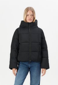 Chaqueta acolchada negra con un alto cuello, cierre de cremallera y diseño acolchado. Presenta un ajuste amplio y mangas largas. Llevada sobre unos jeans azules.