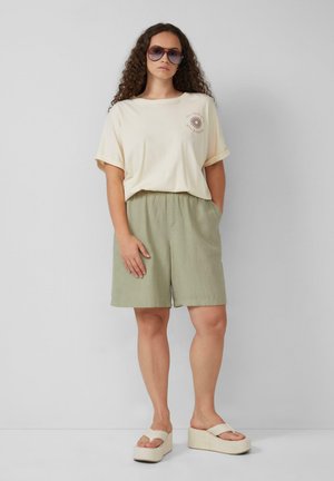 T-shirt beige à manches courtes avec un graphisme de soleil, associé à un short vert olive. La tenue est complétée par des sandales à plateformes de couleur crème.