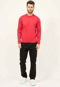 Maglione rosso a maniche lunghe con scollatura tonda e piccolo logo embossato sul petto, abbinato a pantaloni neri e sneakers bianche.