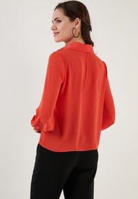 Red, long-sleeve blouse with a collar, smooth texture, and subtle puffed sleeves. Paired with black pants, viewed from the back.