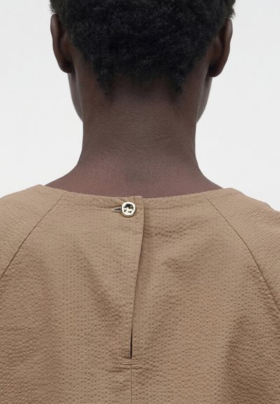 Άτομο με κοντά σγουρά μαλλιά φοράει μπλούζα μπεζ με υφή, στερεωμένη πίσω με ένα μικρό χρυσό κουμπί κοντά στον λαιμό.