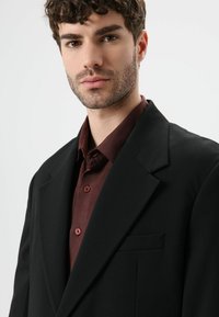 Zwarte blazer met schoudervulling en een inkeping in de revers, een enkele zak, gedragen over een bordeauxrode blouse met rode knopen. Gladde stof met een gestructureerde pasvorm.