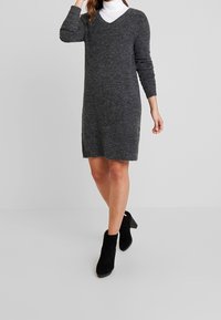 Robe-pull grise en maille avec un col en V, des manches longues et une coupe droite, associée à des bottines en suède noir et un col roulé en dessous.