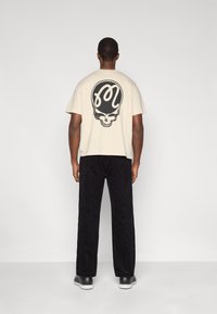 T-shirt en coton beige avec un motif de crâne noir dans le dos. Associé à un pantalon en velours côtelé noir, le modèle pose devant un fond neutre.