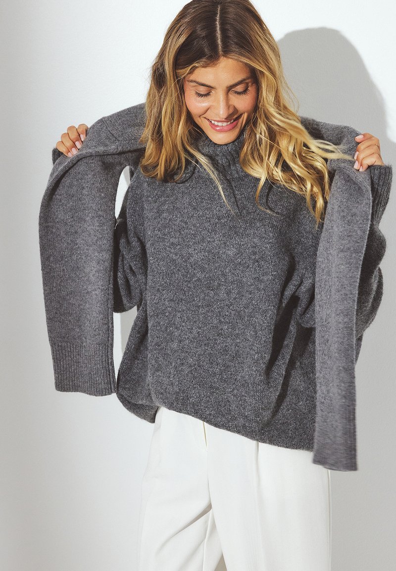 Grijze oversized trui met geribbelde manchetten en zoom, gemaakt van zachte stof, met een relaxte pasvorm en verlaagde schouders, gedragen over lichtgekleurde broek.