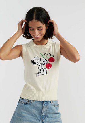 Femme souriante ajuste ses cheveux en portant un pull sans manches blanc avec Snoopy et deux cerises avec le texte « so sweet », associé à un jean bleu.