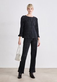 Ensemble deux pièces noir à motifs, composé d'un haut à manches volantes et d'un pantalon évasé. Le tissu présente un design texturé et une finition douce. Accessoirisé avec un sac à main.