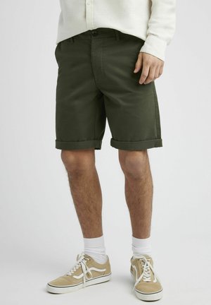 Shorts en coton vert olive avec ourlets repliés, poches latérales et une seule fermeture à bouton, associés à des baskets beiges et des chaussettes blanches.