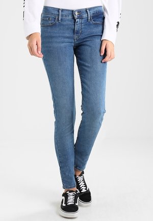 Niebieskie jeansy typu slim fit noszone z czarno-białymi trampkami, połączone z białą bluzką z długim rękawem.