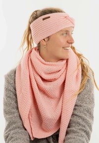 Femme souriante, portant un bandeau en tricot rose et une écharpe assortie sur un pull gris texturé, regardant vers sa droite contre un fond blanc.