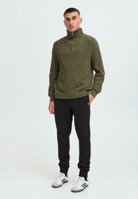 Olive green ribbed pullover with a high collar, paired with black sweatpants. Footwear features white base with black and gray accents.
