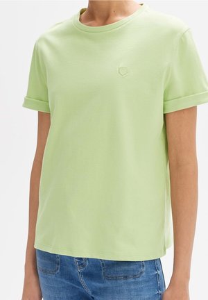 Osoba nosząca jasnobeżową koszulę z krótkim rękawem z podwiniętymi mankietami i małym haftowanym serduszkiem na lewej piersi, w połączeniu z niebieskimi dżinsami.