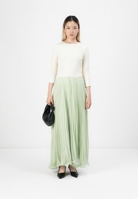 Cremfarbenes Langarmoberteil, kombiniert mit einem fließenden, plissierten mintgrünen Rock. Eine schwarze Handtasche in der Hand und spitze schwarze Ballerinas runden das Outfit ab.