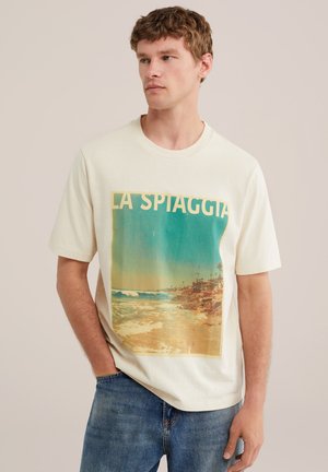 Jeune homme aux cheveux courts et bouclés portant un t-shirt beige avec une scène de plage vintage et le texte "LA SPIAGGIA", associé à un jean bleu.