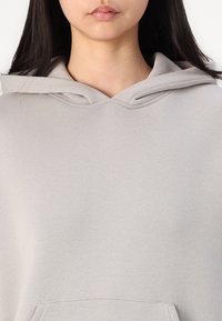 Hoodie gris clair avec une poche kangourou, capuche pliée et tissu doux et texturé. Design minimaliste sans motifs ni accents visibles.
