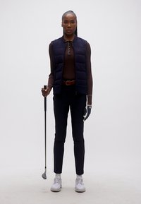 Γυναίκα που φοράει γάντι γκολφ και γιλέκο κρατώντας μπαστούνι γκολφ, σε λευκό φόντο.