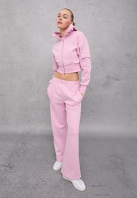 Felpa cropped rosa con zip e tasche, abbinata a pantaloni della tuta rosa larghi. Sneakers bianche completano l'outfit. Texture liscia in tutto.