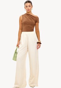 Bruin een-schouder bodysuit gecombineerd met witte geplooide brede broek, met een groene handtas en gestapelde armbanden. Glad textuur.