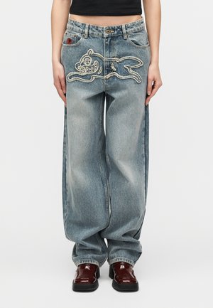 Ženska, oblečena v ohlapne izprane modre kavbojke z našitim obrisom risanega zajčka na sprednji strani, kombinirane s temno rjavimi čevlji.