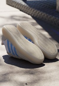 Par hvide sneakers med lyseblå striber, sålerne opad, placeret på solbelyst beton nær rebbarriere.