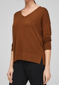 Brun V-hals tröja med rullade ärmar, sidslitsar vid fållen, och en mjuk textur. Tillverkad av ett lätt stickat tyg.