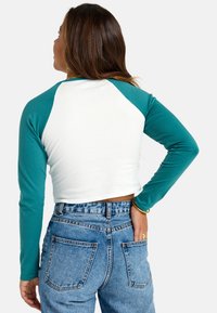 Langärmeliges, kurzes Shirt mit türkisfarbenen Ärmeln und weißem Körper, kombiniert mit hoch geschnittenen Jeans aus Denim, die Gesäßtaschen und eine helle Waschung aufweisen.