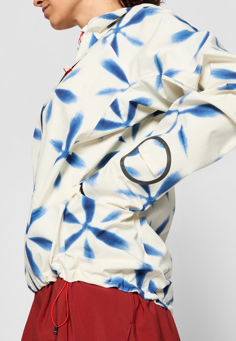 Weiße Jacke mit blauem Blumenmuster, ausgestattet mit einem elastischen Saum, rotem Kordelzug und einem schwarzen kreisförmigen Detail am Ärmel.