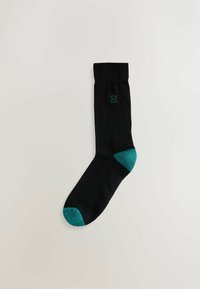 Schwarze Socke mit dunklem Türkis an Ferse und Zehen, hergestellt aus einem glatten Material, mit einem kleinen Logo nahe dem oberen Rand.