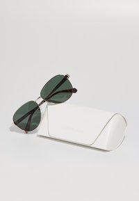 Lunettes de soleil teintées en vert avec des verres triangulaires et des accents en métal doré, reposant sur un étui rigide blanc et texturé. L'étui a un design lisse et courbé.