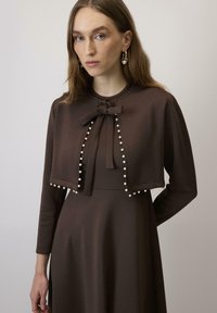 Rochie maro cu o capa scurta, decorata cu o fundita si accente de perle pe margini. Maneci lungi si un material texturat, neted.