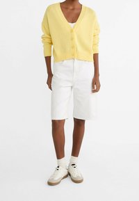 Yellow knitted cardigan with a v-neck and six buttons, paired with white knee-length shorts and cream sneakers with brown accents.