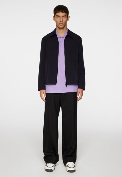 Giacca navy con colletto, indossata sopra una polo di colore viola chiaro. Pantaloni neri larghi e sneakers multicolori completano l'outfit.