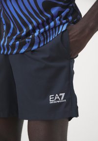Pantalones cortos deportivos de color navy con bolsillos laterales, que presentan un logo blanco en contraste y una tela suave y ligera. Parte superior azul con patrón que se superpone en la cintura.