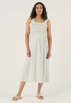 Woman with shoulder-length curly hair wearing a sleeveless light plaid midi dress and slip-on sandals, standing against a plain background.