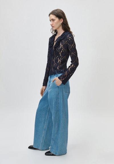 Kobieta ubrana w granatową koronkową bluzkę z falbankami, wpuszczoną w wysokie, szerokie dżinsy o kroju dzwonów oraz czarne buty, stojąca bokiem.