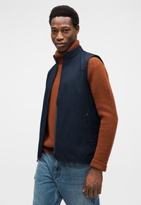 Μπλε σκούρο αμάνικο μπουφάν με ψηλό γιακά, φορεμένο πάνω από ένα σκουροκόκκινο ριγωτό ζιβάγκο. Το τζιν παντελόνι έχει άνετη εφαρμογή.