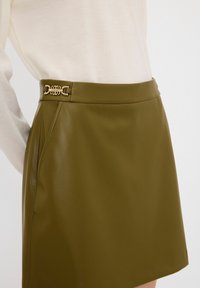 Mini gonna in eco-pelle verde oliva con una texture liscia, dotata di una fibbia decorativa in tono oro in vita e tasche laterali.