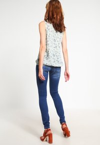 Haut sans manches floral bleu clair, associé à un jean skinny bleu foncé et des sandales à talons ouverts marron. Vue de dos, tissu lisse et coupe ajustée.