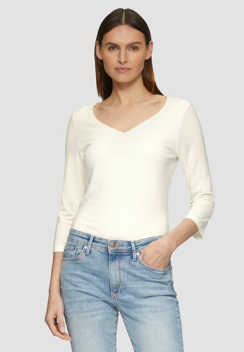 Witte blouse met lange mouwen en een V-hals ontwerp, met een gladde textuur, gecombineerd met lichtblauwe jeans. De stof lijkt zacht en geschikt voor casual wear.