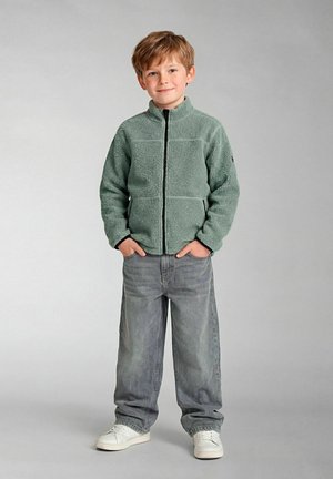 Mlad fant stoji z rokami v žepih, oblečen v svetlo zelen pulover z zadrgo, široke sive kavbojke in bele superge, na enobarvnem ozadju.