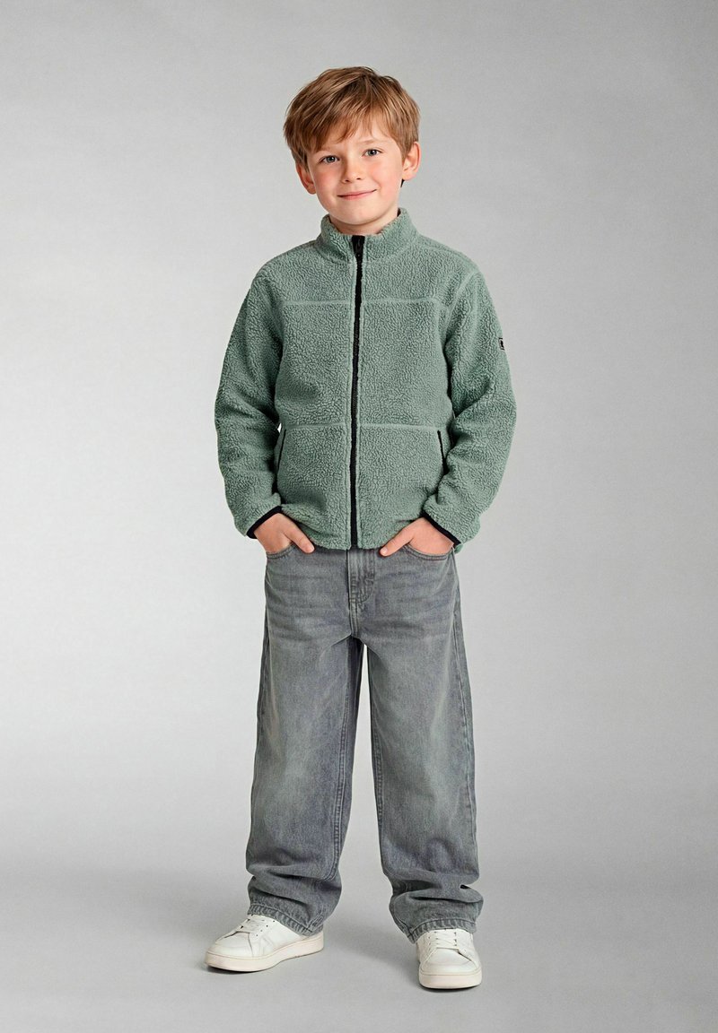 Jonge jongen staat met handen in zakken, draagt een lichtgroene fleece met rits, losse grijze spijkerbroek en witte sneakers tegen een effen achtergrond.