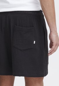 Pantalones cortos de algodón negros con un patrón texturizado, que cuentan con una cintura elástica, bolsillos laterales y una pequeña etiqueta de marca en el bolsillo trasero.
