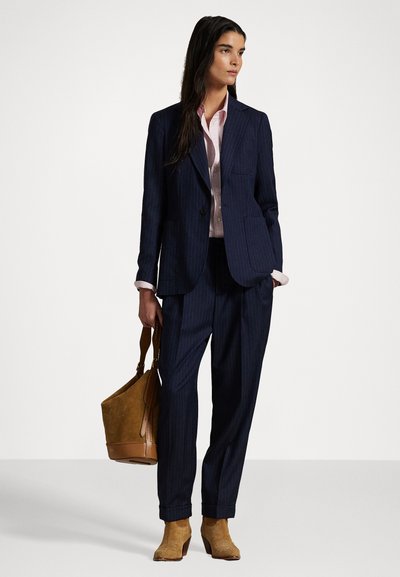 Navy pinstriped suit with a tailored blazer and loose trousers. Light pink button-up shirt underneath. Brown suede ankle boots and a tan handbag.